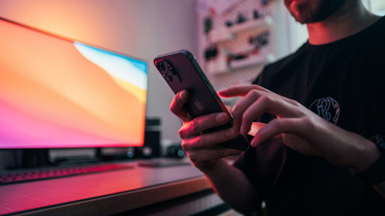 A man in front of a monitor looking down at his smartphone.