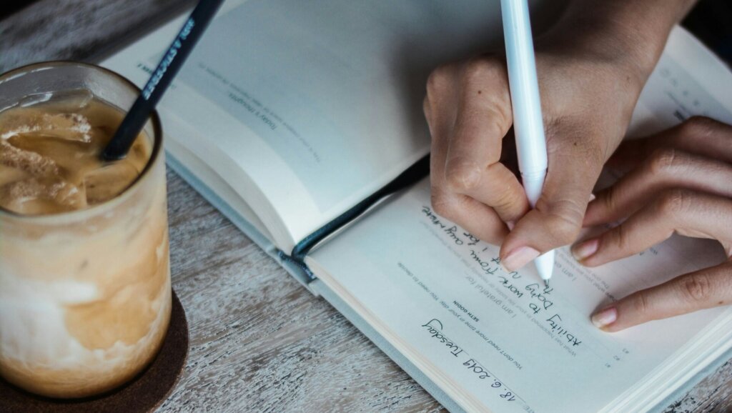 Closeup of a hand, writing in a journal. A drink positioned to the left.