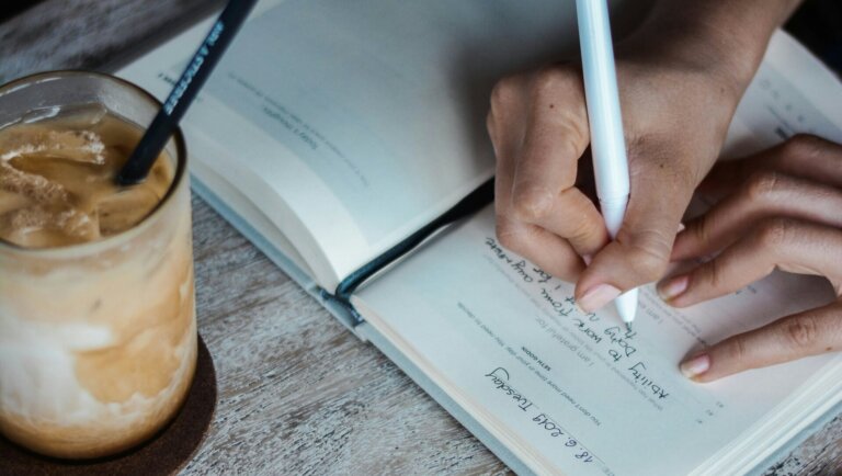 Closeup of a hand, writing in a journal. A drink positioned to the left.