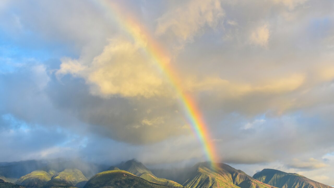 An image of a rainbow in a cloudy sky above some green hills.